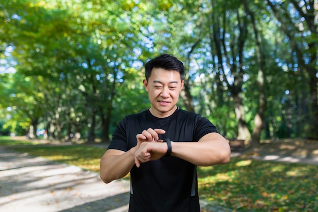 Homem asiático sorridente e bem-sucedido correndo e se exercitando no parque, olhando para o relógio inteligente e