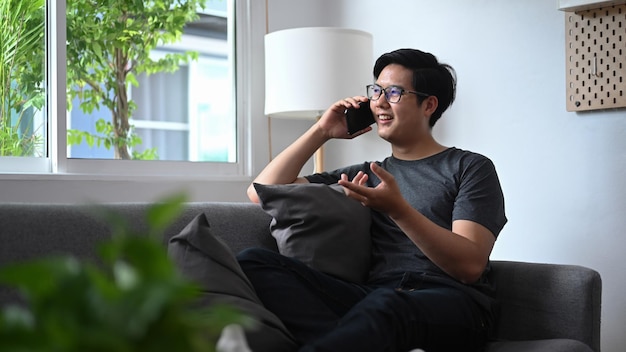 Homem asiático sorridente falando no telefone inteligente e sentado no sofá na sala de estar