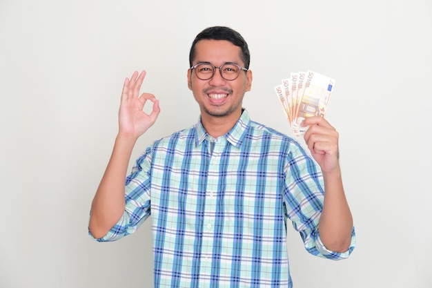 Homem asiático sorrindo feliz e dando sinal de dedo OK enquanto segura a moeda Euro