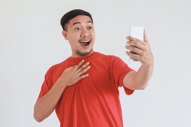 Homem asiático surpreso e chocado com camiseta vermelha usando smartphone em fundo branco