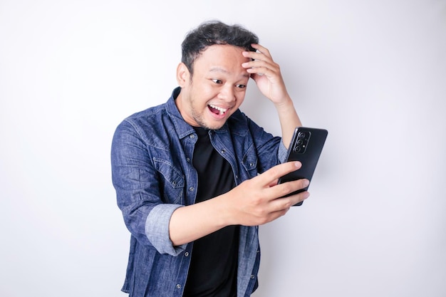 Homem asiático surpreso vestindo camisa azul marinho apontando para seu smartphone isolado por fundo branco