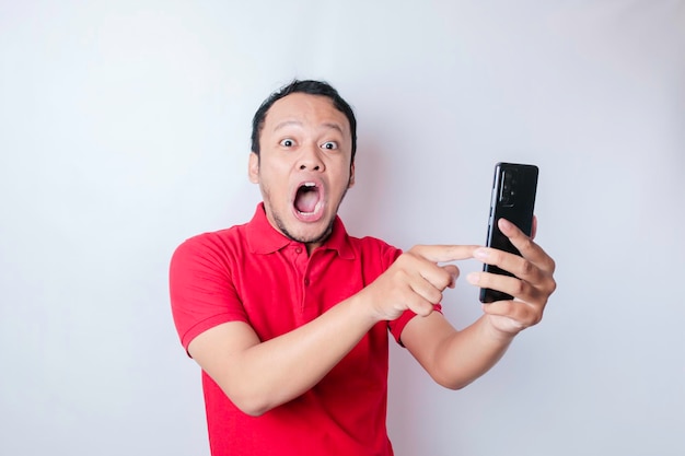Homem asiático surpreso vestindo camiseta vermelha apontando para seu smartphone isolado por fundo branco