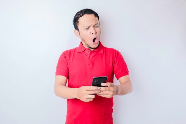 Homem asiático surpreso vestindo camiseta vermelha apontando para seu smartphone isolado por fundo branco