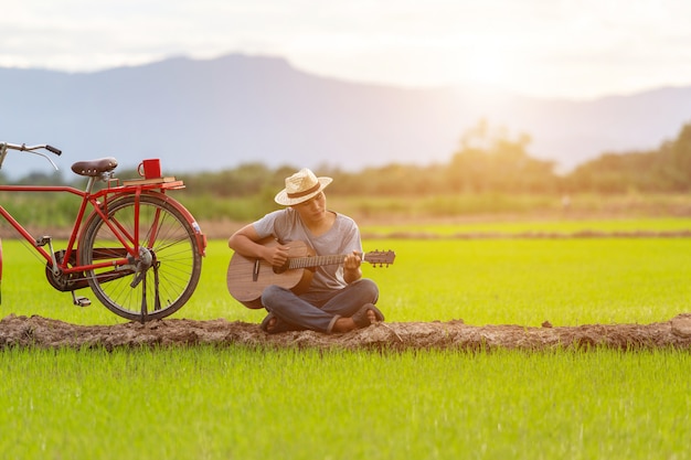 Homem asiático tocando guitarra no campo de arroz verde na hora do sol