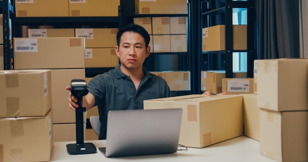 Foto homem asiático usa dados de leitura de código de barras na caixa e digita registrar pedido on-line no laptop no armazém