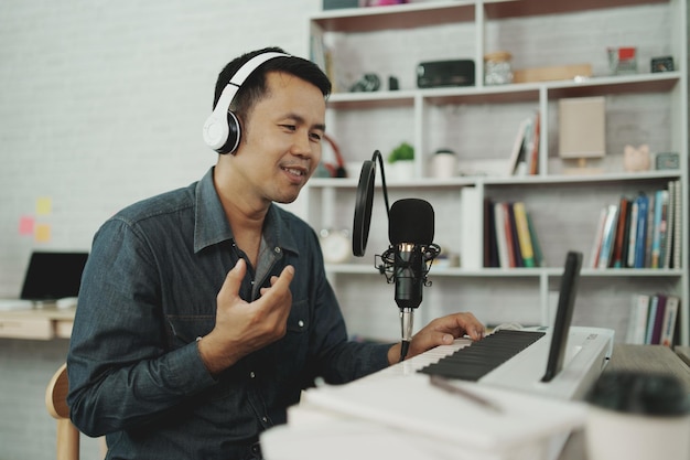 Homem asiático usando fones de ouvido brancos toca piano e canta uma música aprendendo on-line com telefone celular ou grava som com um microfone e usa programa de gravação de música engenheiro de som grava música