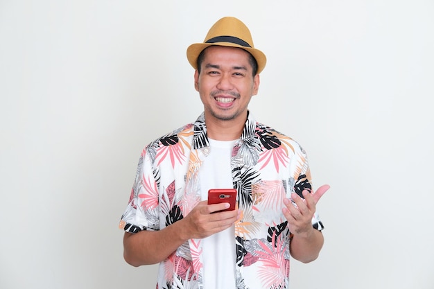 Homem asiático vestindo camisa de praia e chapéu sorrindo feliz enquanto segura o telefone