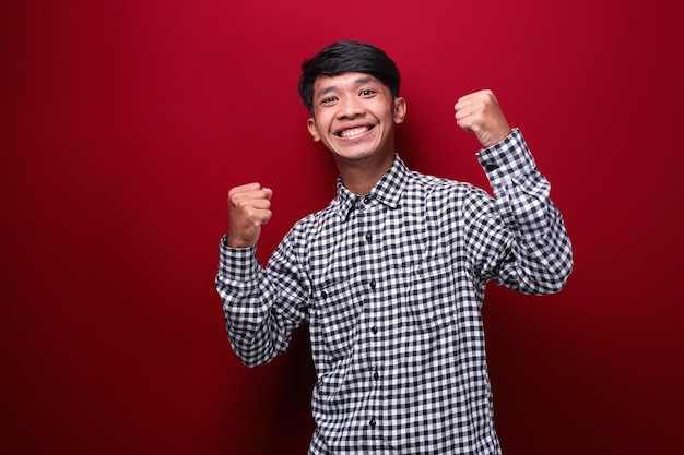Homem asiático vestindo camisa xadrez com expressão feliz mostrando vitória em fundo vermelho
