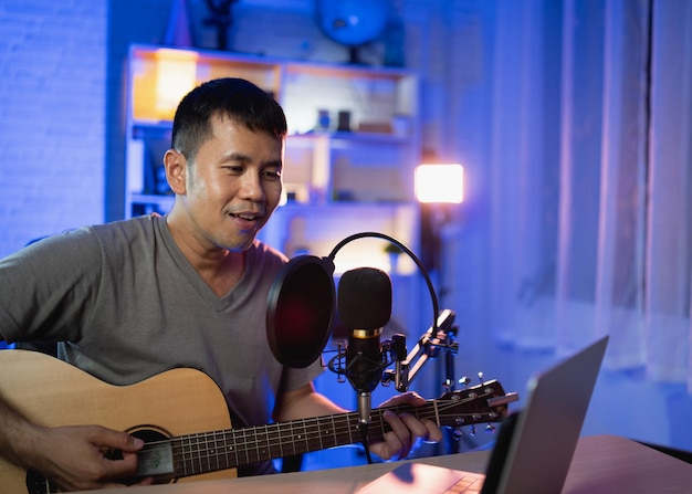 Homem asiático youtuber ao vivo transmitindo performance tocando violão e cantando uma música Homem asiático ensinando violão e cantando online Músico gravando música com laptop e tocando violão