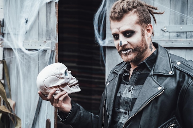 Homem assustador com dreadlocks olhando para o crânio Celebrando o halloween Terrível maquiagem de monstro de rosto branco preto e imagem de fantasia eleganteHorrordiversão na festa infantil no celeiro na ruaCamisajaquetas