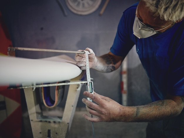 Homem atento com cabelos loiros e tatuagens nos braços em roupas casuais e respirador medindo a espessura da prancha de surf com pinça durante o trabalho na carpintaria