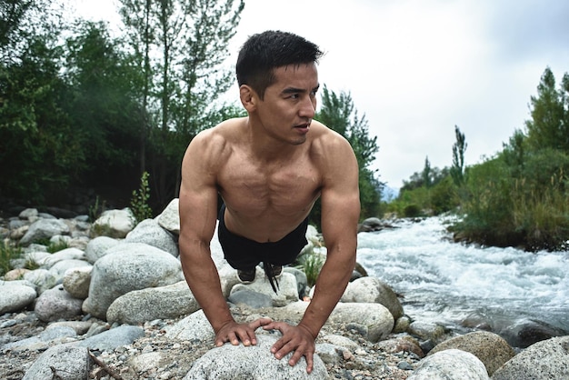 Homem atleta muscular cazaque treina e exercícios à beira do rio na natureza. Bonito asiático faz exercícios físicos extremos ao ar livre