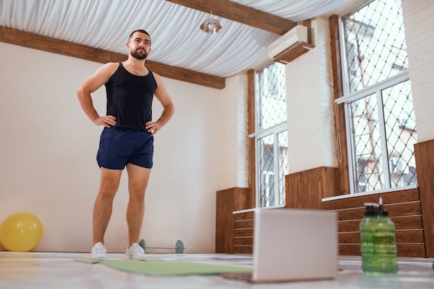 Homem atleta treina duro no ginásio vazio ou em pé na frente do laptop em casa. Aquecimento de jovem auto-isolado e motivado, fazendo exercícios especiais para os músculos. Exercite-se durante o bloqueio do coronavírus.