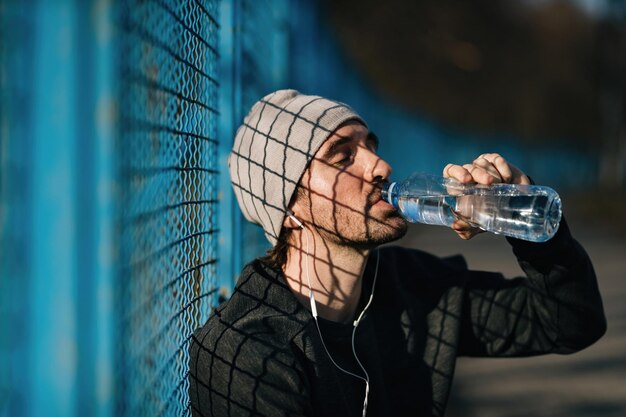 Homem atlético cansado bebendo água com os olhos fechados enquanto faz uma pausa no exercício ao ar livre.