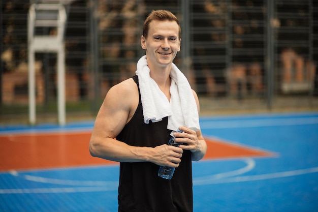 Homem atlético com toalha nos ombros bebendo água e relaxando após um treino intenso ao ar livre