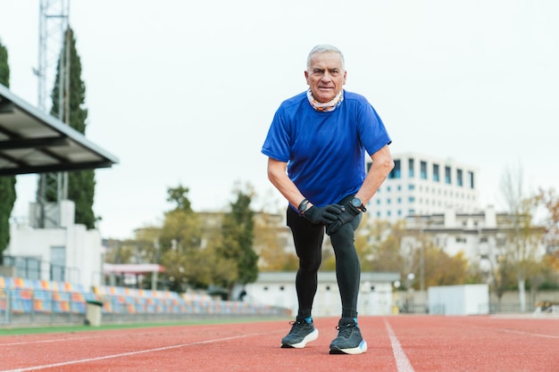 Homem atlético idoso faz uma pausa para descansar no campo de atletismo vestido com trajes de corrida mostrando determinação