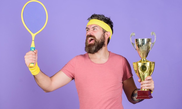 Homem atlético segura raquete de tênis e taça de ouro Jogador de tênis ganha campeonato Homem barbudo atleta de sucesso Primeiro lugar Conquista esportiva Campeão de tênis Vença o jogo de tênis Comemore a vitória