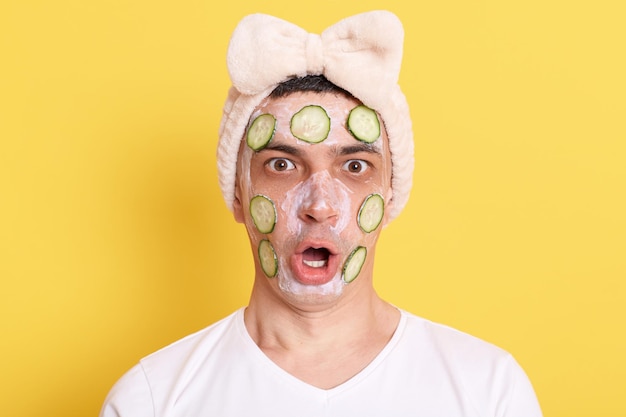 Homem atônito chocado na camiseta branca e faixa de cabelo com laço posando com máscara facial e pepino isolado sobre fundo amarelo expressão de surpresa olhando para a câmera com olhos grandes e boca aberta