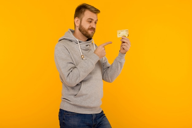 Homem atraente com um moletom cinza aponta um dedo para o cartão de crédito que está segurando em um fundo amarelo