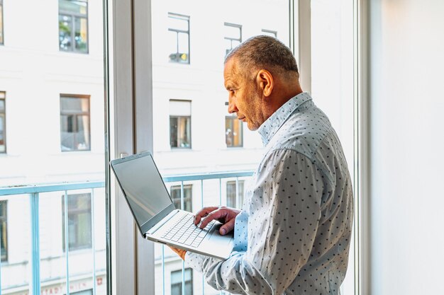 Homem atraente de camisa em pé perto da janela com laptop