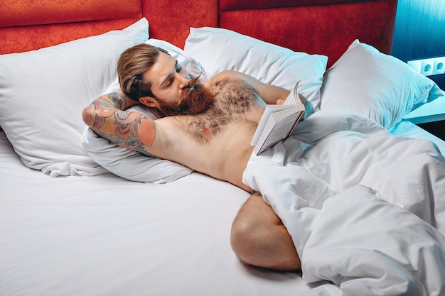 Homem atraente nu sexy com bigode e barba deitado em sua cama branca e lendo um livro. Conceito noturno