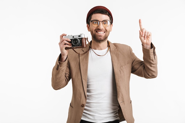 Homem atraente usando um chapéu moderno, regozijando-se ao segurar e fotografar na frente retrô isolada sobre uma parede branca