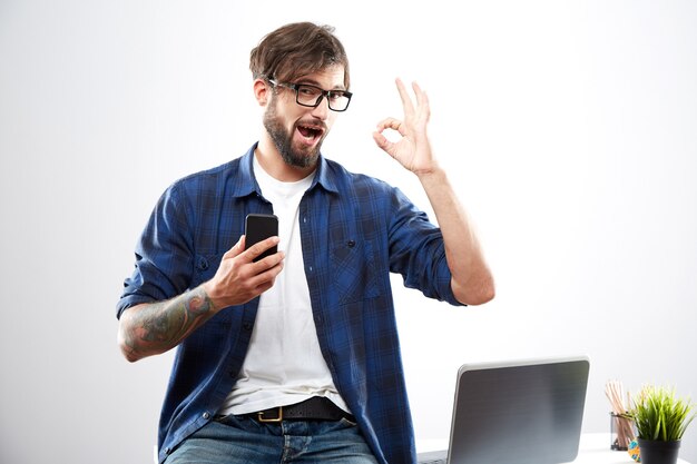 Homem atraente, vestindo camisa azul e óculos, sentado com o laptop, conceito freelance, retrato, trabalho online, mostrando sinal de OK.