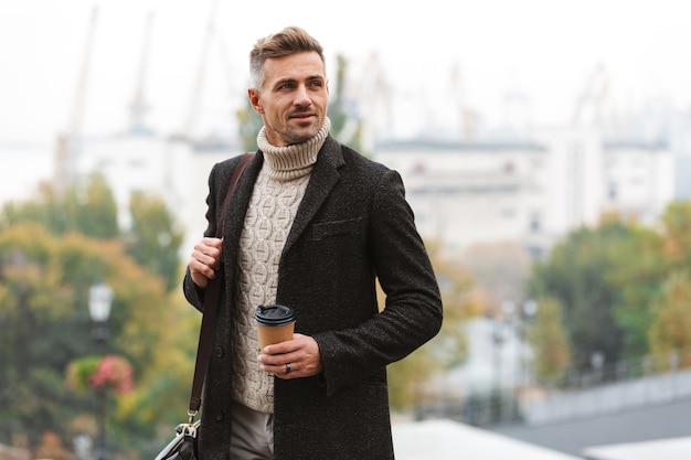 Homem atraente vestindo jaqueta caminhando ao ar livre, segurando uma xícara de café para viagem