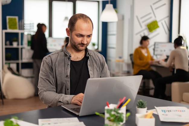 Homem autêntico gerente sentado à mesa em um escritório de inicialização moderno, enviando e-mails digitando no laptop