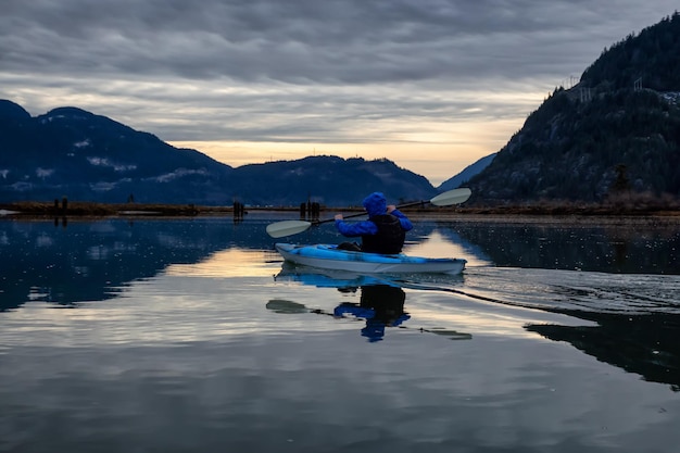 Homem aventureiro andando de caiaque em águas tranquilas durante um pôr do sol nublado de inverno