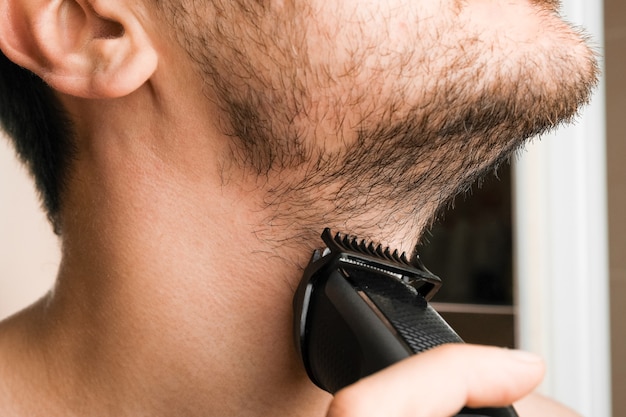 Homem barbeando a barba e o pescoço com um barbeador elétrico. Rotina matinal. Aparar a barba com um aparador.