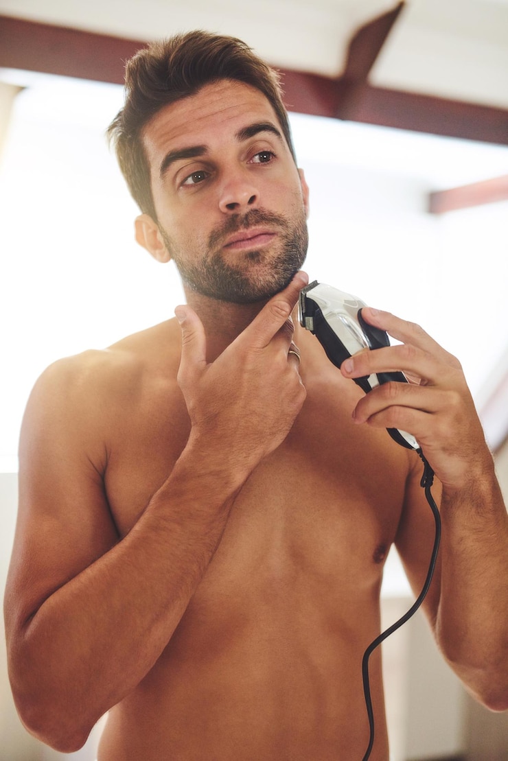 Foto homem barbeando e rosto com barbeador elétrico para remoção de pêlos cuidados estéticos ou limpeza de beleza em casa cara sem camisa barbear barba com aparador de lâmina e ferramenta cosmética no banheiro pela manhã