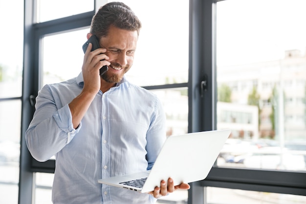 Homem barbudo alegre no escritório, falando pelo telefone celular.