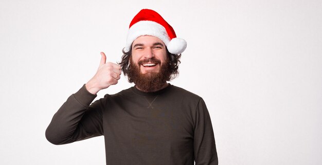 Homem barbudo alegre usando chapéu de Papai Noel e mostrando o polegar