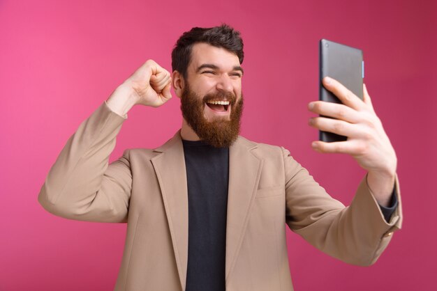 Homem barbudo animado está olhando para seu tablet ou fazendo um selfie enquanto mostra o gesto de vencedor.