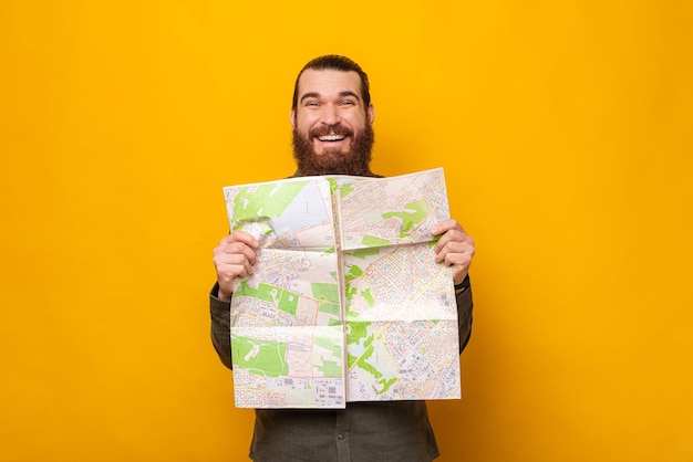Homem barbudo animado está segurando um mapa de papel enquanto sorri para a câmera Studio atirou sobre fundo amarelo