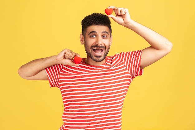 Homem barbudo animado positivo em camiseta listrada brincando com corações de brinquedo vermelho, mostrando seu carinho e devoção, romance. Tiro de estúdio interior isolado em fundo amarelo