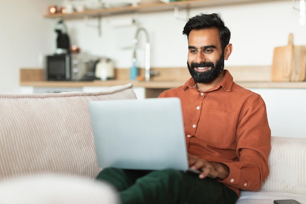 Homem barbudo árabe feliz a surfar na web no computador em casa.