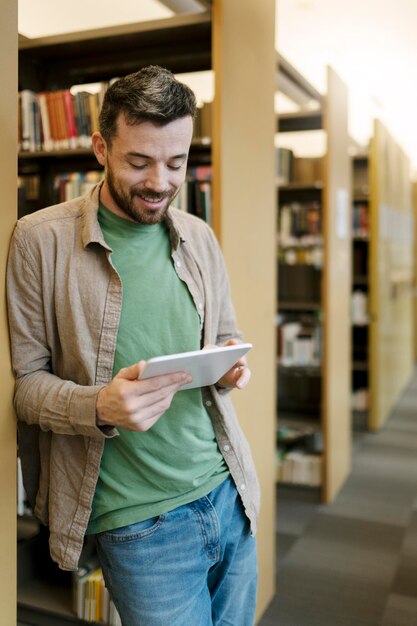 Homem barbudo atraente está parado perto da estante de livros usando o livro de busca de tablet