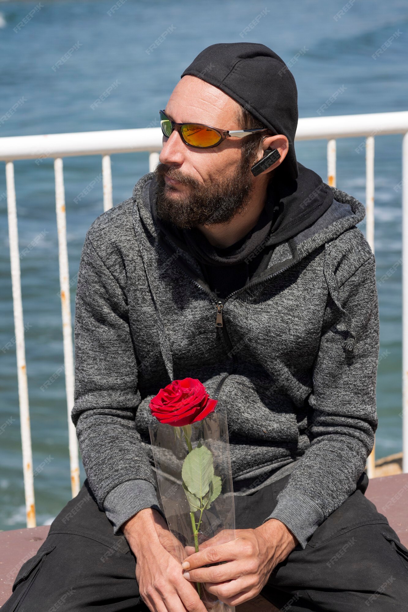 Homem barbudo bonito americano usando óculos escuros, boné, jaqueta com  capuz com rosa esperando sua mulher na praia do mar. homem russo caucasiano  brutal de moda olha para o lado, pose relaxada