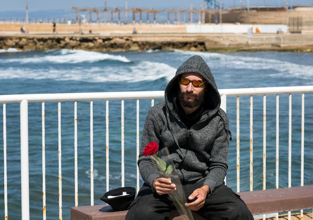 Homem barbudo bonito americano usando óculos escuros e jaqueta com capuz contra o mar azul em um dia ensolarado. Cara russo caucasiano brutal segurando uma flor rosa vermelha na mão. Feriado romântico e dia dos namorados