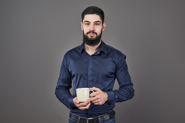 Homem barbudo bonito com barba de cabelo elegante e bigode no rosto sério na camisa segurando copo branco ou caneca bebendo chá ou café no estúdio em cinza