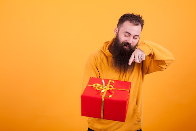 Homem barbudo bonito com capuz amarelo apontando na caixa de presente vermelha sobre fundo amarelo. Homem bonito. Presente surpresa.