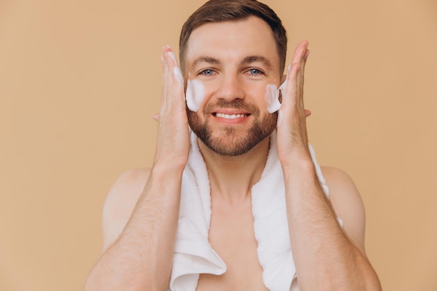 Homem barbudo bonito e feliz com uma toalha ao redor do pescoço aplicando espuma de lavagem facial em fundo bege conceito de beleza e cuidado