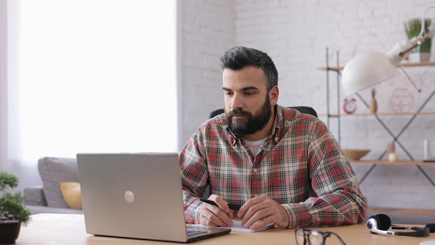 Homem barbudo bonito ensinando on-line, falando com a câmera da web do laptop. Educação online, trabalho remoto.