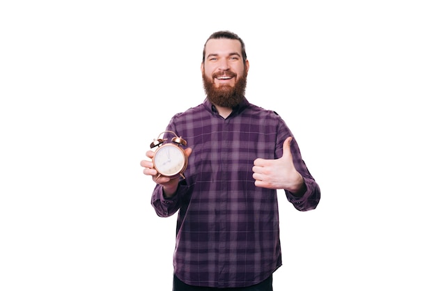 Homem barbudo bonito feliz alegre mostrando o polegar e o despertador