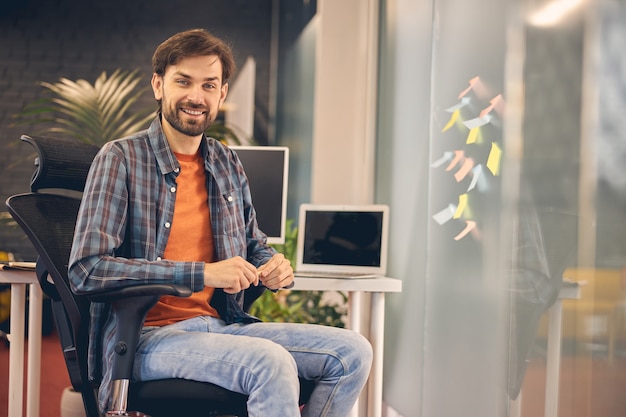 Homem barbudo bonito olhando para a câmera e sorrindo enquanto trabalha em um escritório moderno