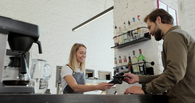 Homem barbudo bonito, pagando por um café para viagem com o celular na cafeteria. Trabalhador que atende um cliente que usa smartphone para pagar na cafeteria.