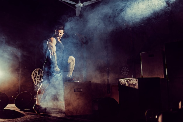 Homem barbudo caucasiano atraente muscular levantando dois kettlebells em um ginásio. Placas de peso, halteres e pneus em segundo plano.