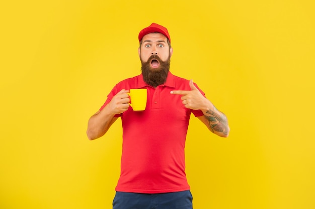 Homem barbudo chocado com boné vermelho casual e camiseta apontando o dedo para a caneca de chá de fundo amarelo